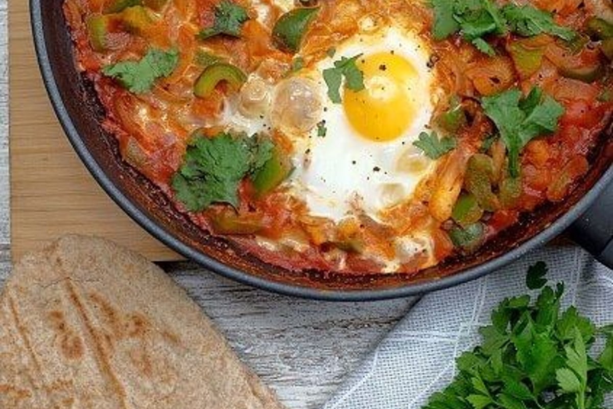 Two eggs cooked in tomato sauce with herbs in a pan, bread, parsley, and pepper bowl