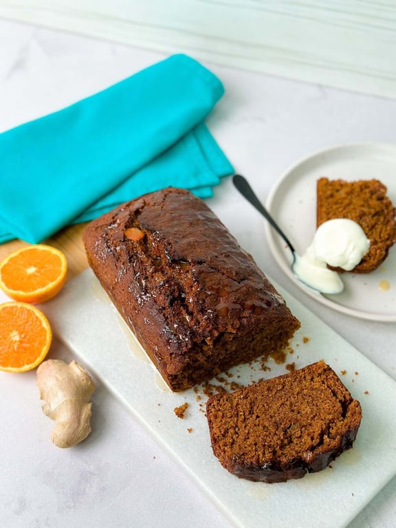 Loaf of gingerbread cake with a slice cut, orange halves, and a piece with cream on a plate