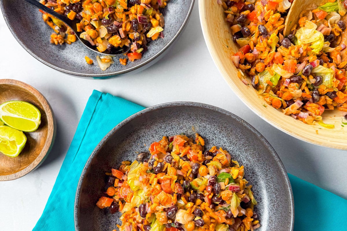 Two bowls and a wooden bowl filled with mixed bean and vegetable salad with lime wedges