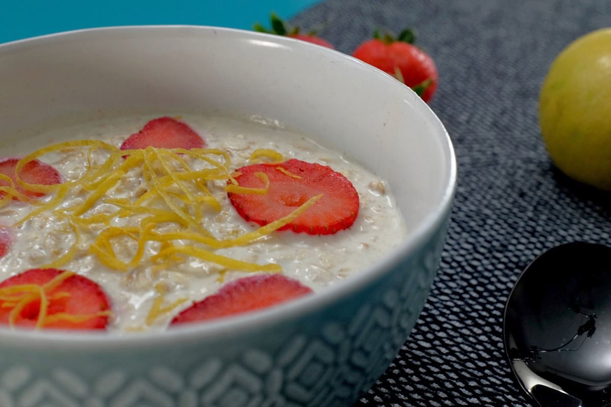 Bowl of oats topped with sliced strawberries and lemon zest beside fresh fruit