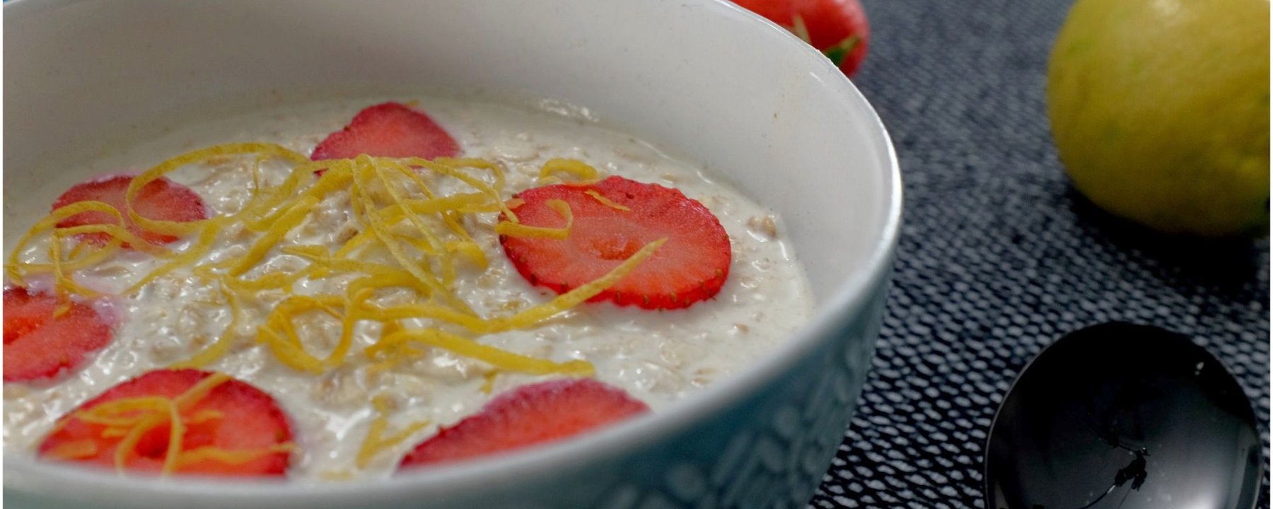 Bowl of oats topped with sliced strawberries and lemon zest beside fresh fruit