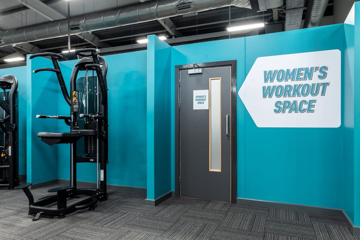 PureGym Birmingham West: Women's workout area with weight machine, teal walls, and sign indicating designated space.