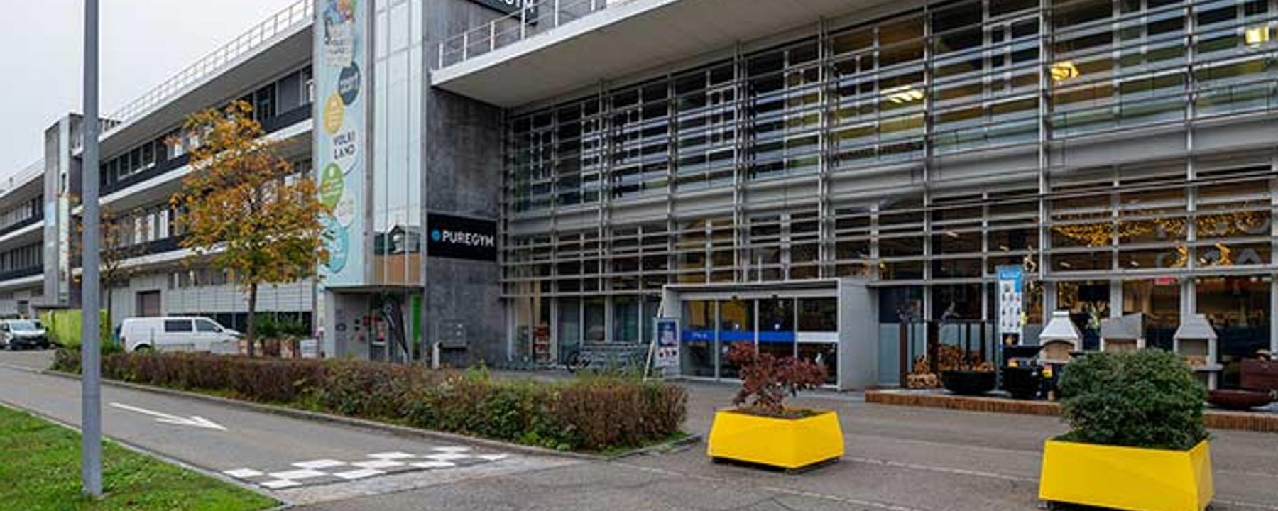 Puregym Volketswil: Grosse Ladenfront mit Parkplatz, Pflanzgefässe im Vordergrund.