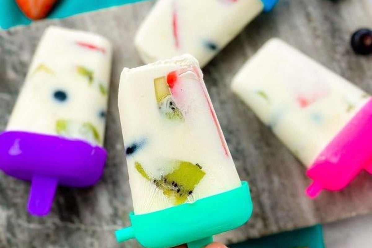 Hand holding a fruit-filled yoghurt popsicle with more on a stone board and scattered frui