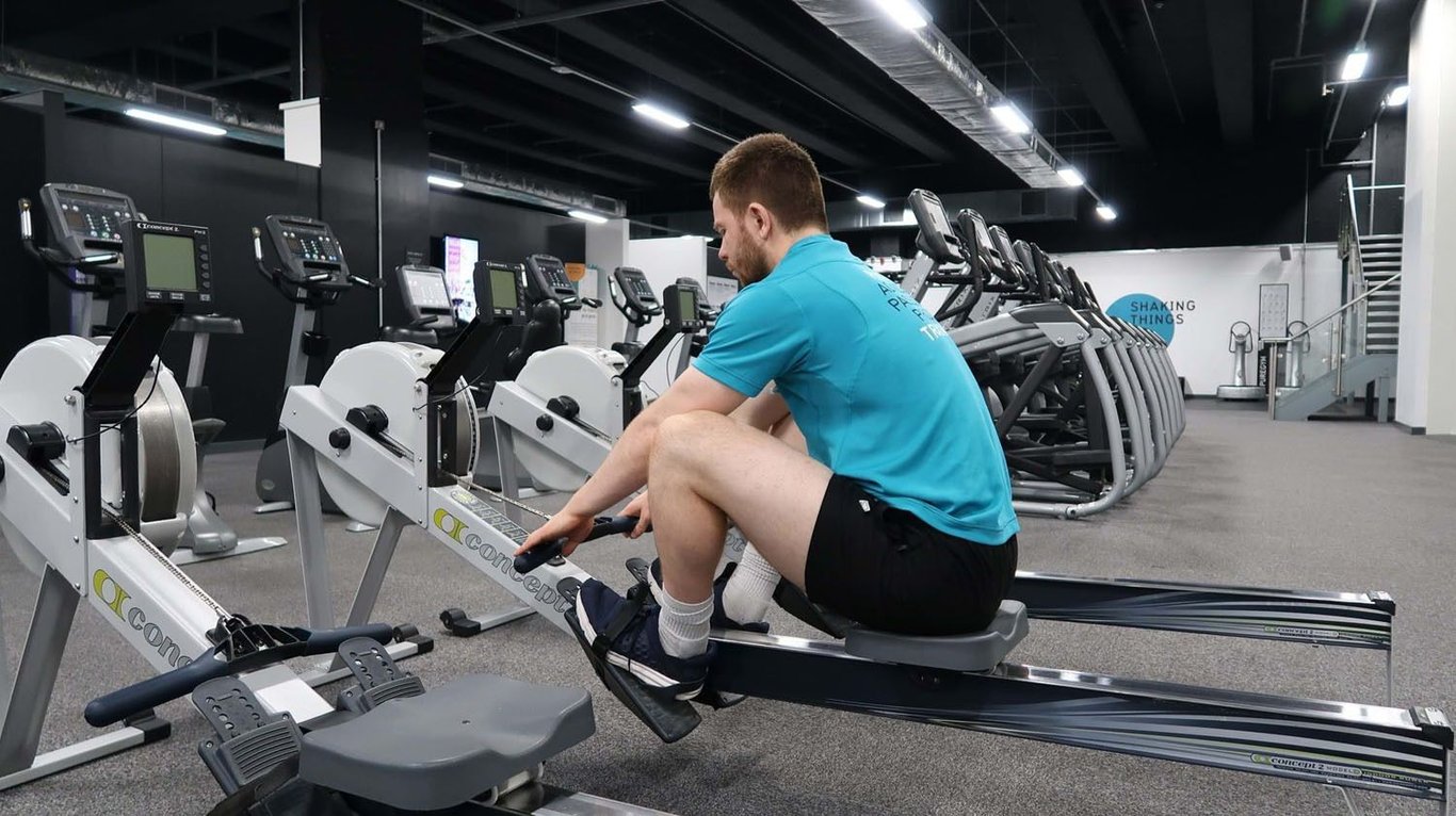 Man in blue shirt using rowing machine in gym with treadmills in background