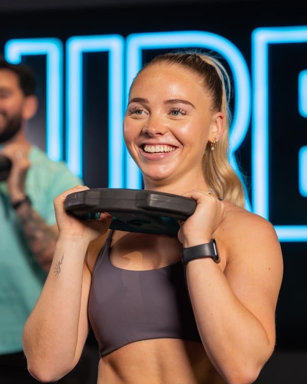 Smiling woman holding a weight plate in a modern PureGym facility with blue neon signage