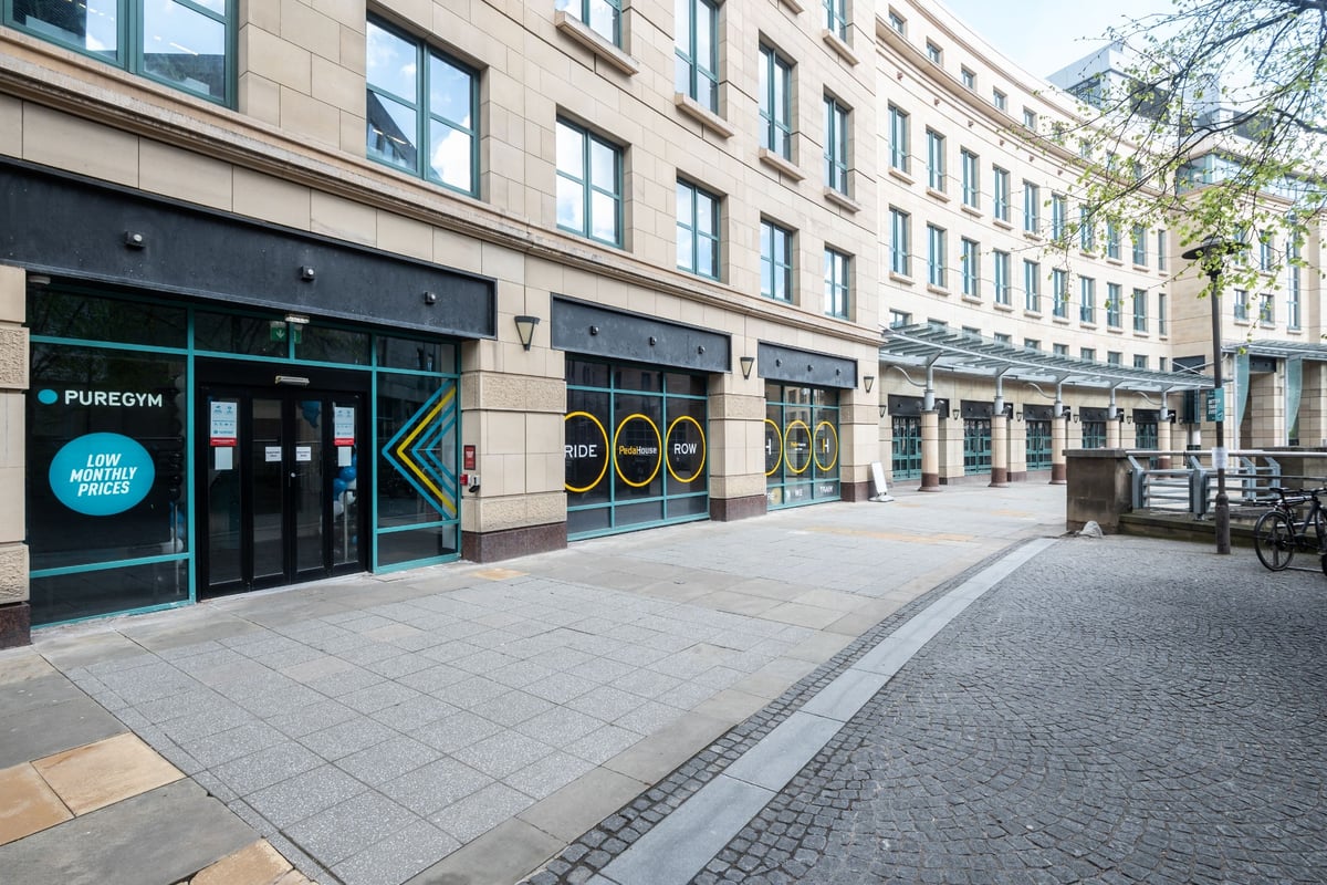PureGym Edinburgh Exchange Crescent: A street with storefront displays, seating, and bikes parked outside a fitness center.
