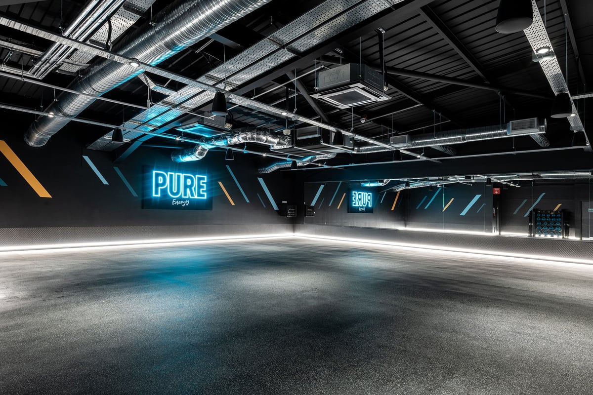 PureGym Cardiff Western Avenue: Exposed ceiling with beams and pipes, neon signage saying "PURE", open concrete floor area.