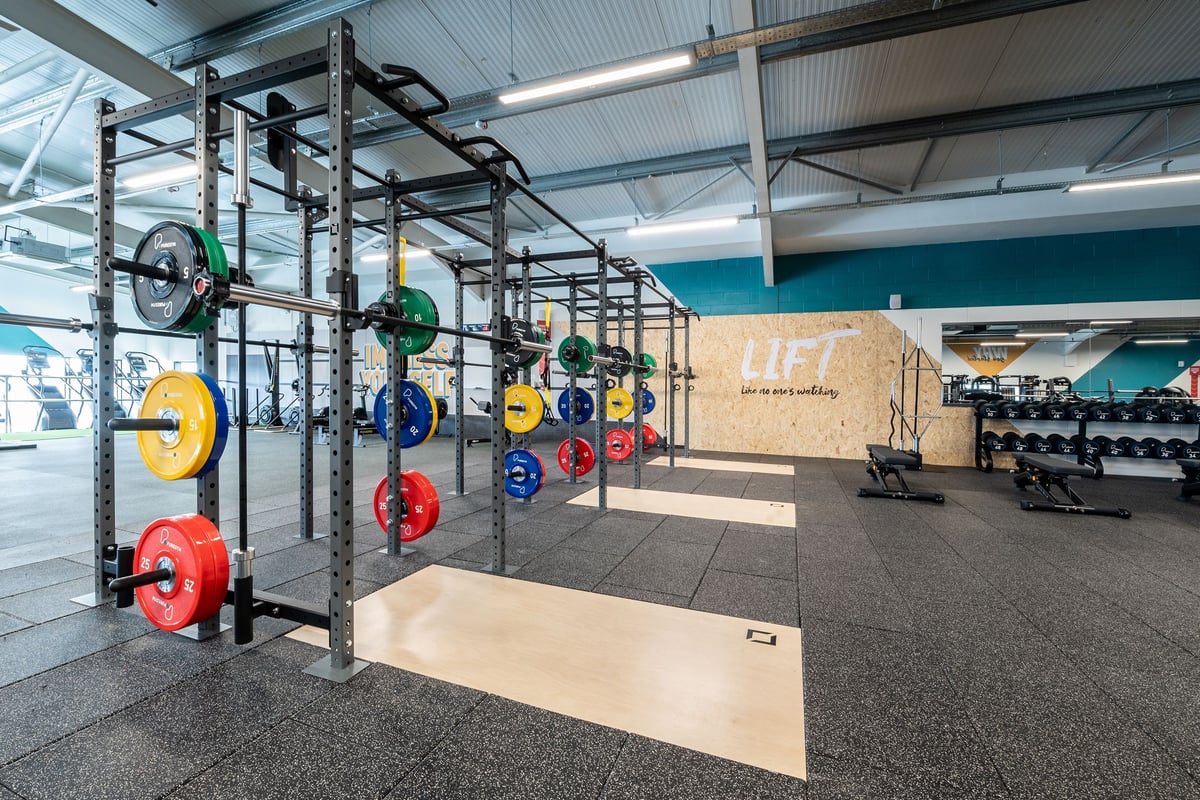 PureGym London Colney: Squat racks, coloured weight plates, lift sign, rubberized flooring in a bright weightlifting area.