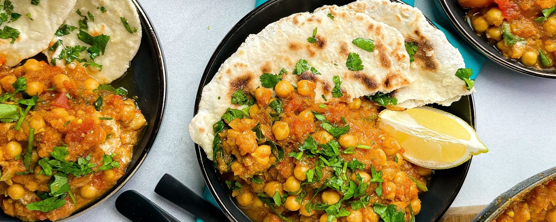 Chickpea curry with fresh coriander served in black bowls with flatbread and lemon wedge