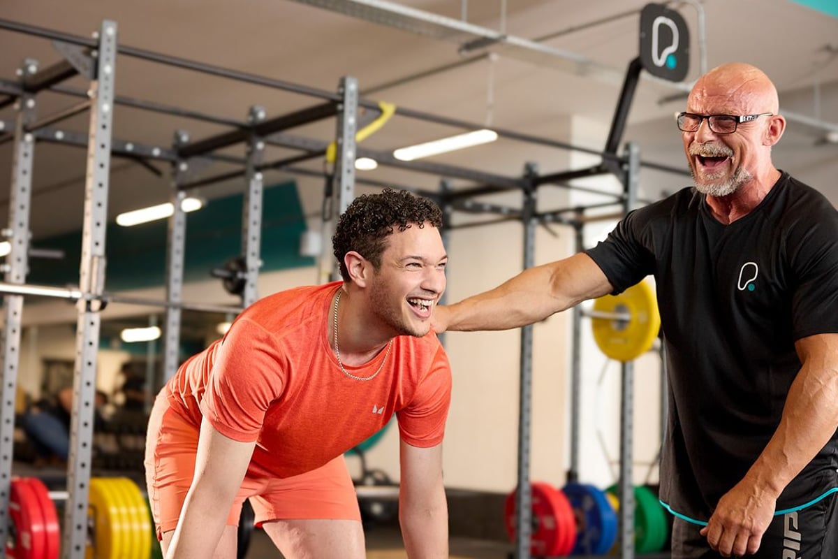 Young man in orange workout clothes laughing with trainer in gym with weights.