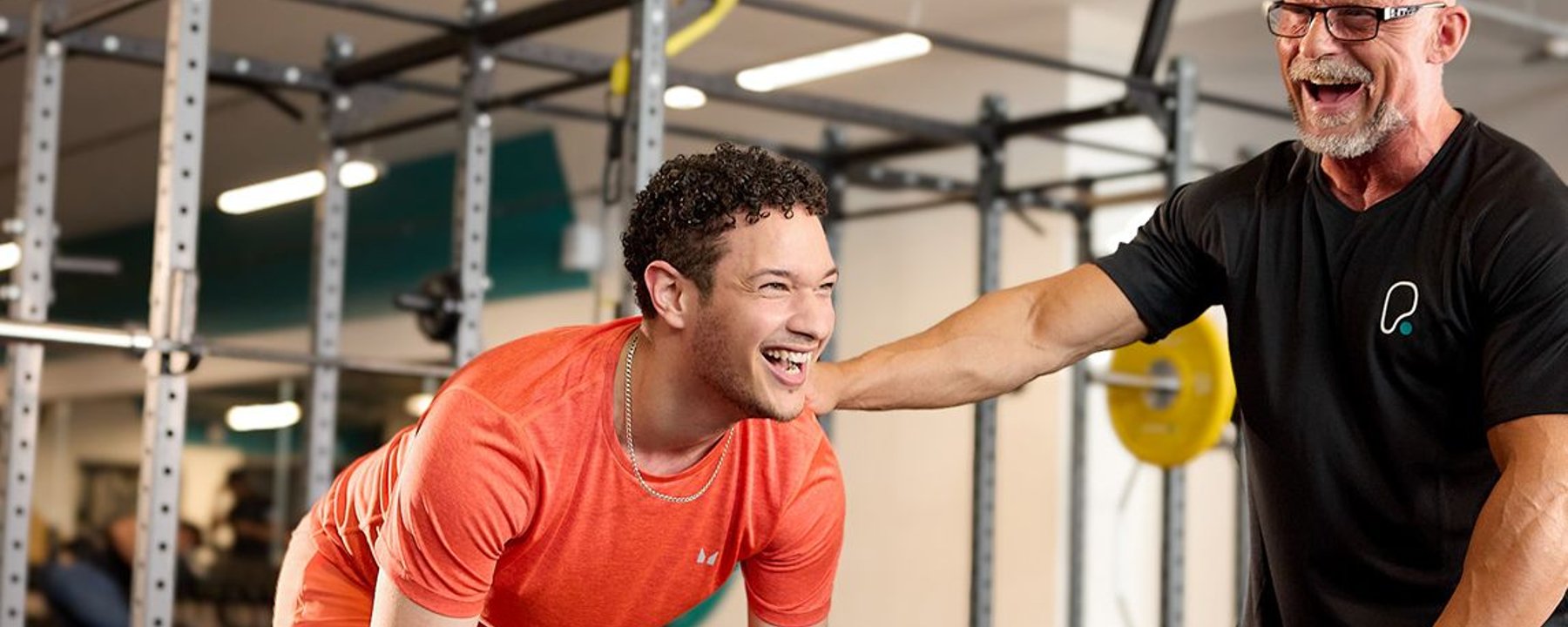 Young man in orange workout clothes laughing with trainer in gym with weights.