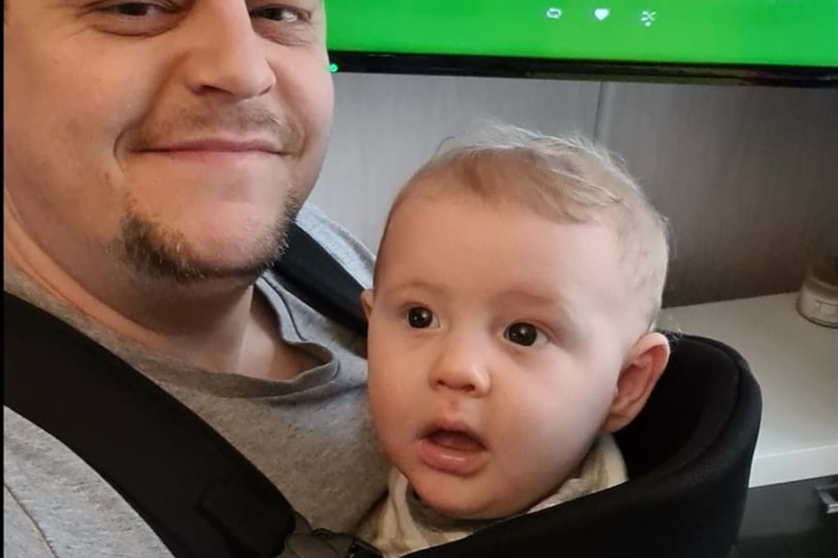 Man smiling with baby in carrier in front of a screen showing music covers and controls