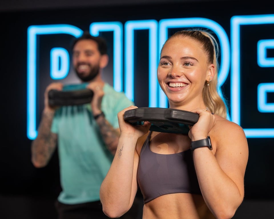 Woman in gym holding weight plate at shoulder height with man exercising behind her