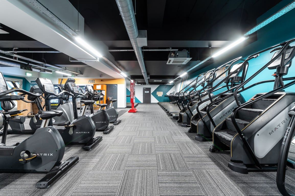 PureGym London Ilford: Rows of elliptical machines and treadmills in a dimly lit gymnasium with blue accent lighting.