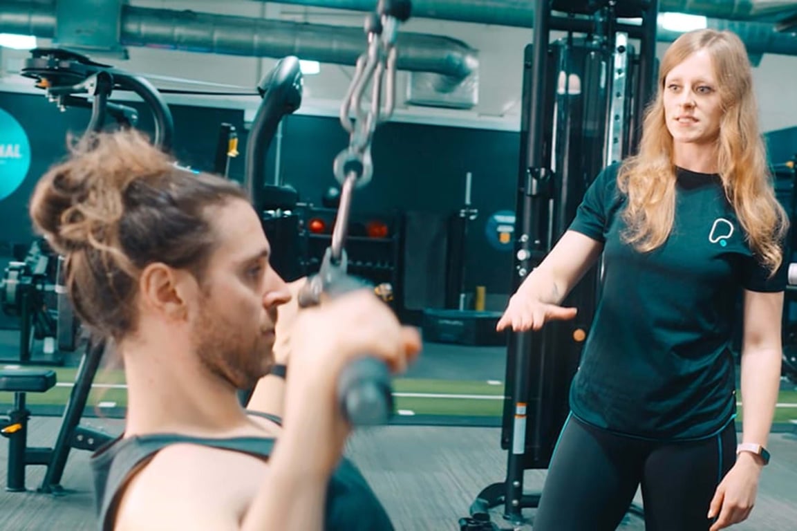 Man lifting weights while woman in sportswear coaches him in gym