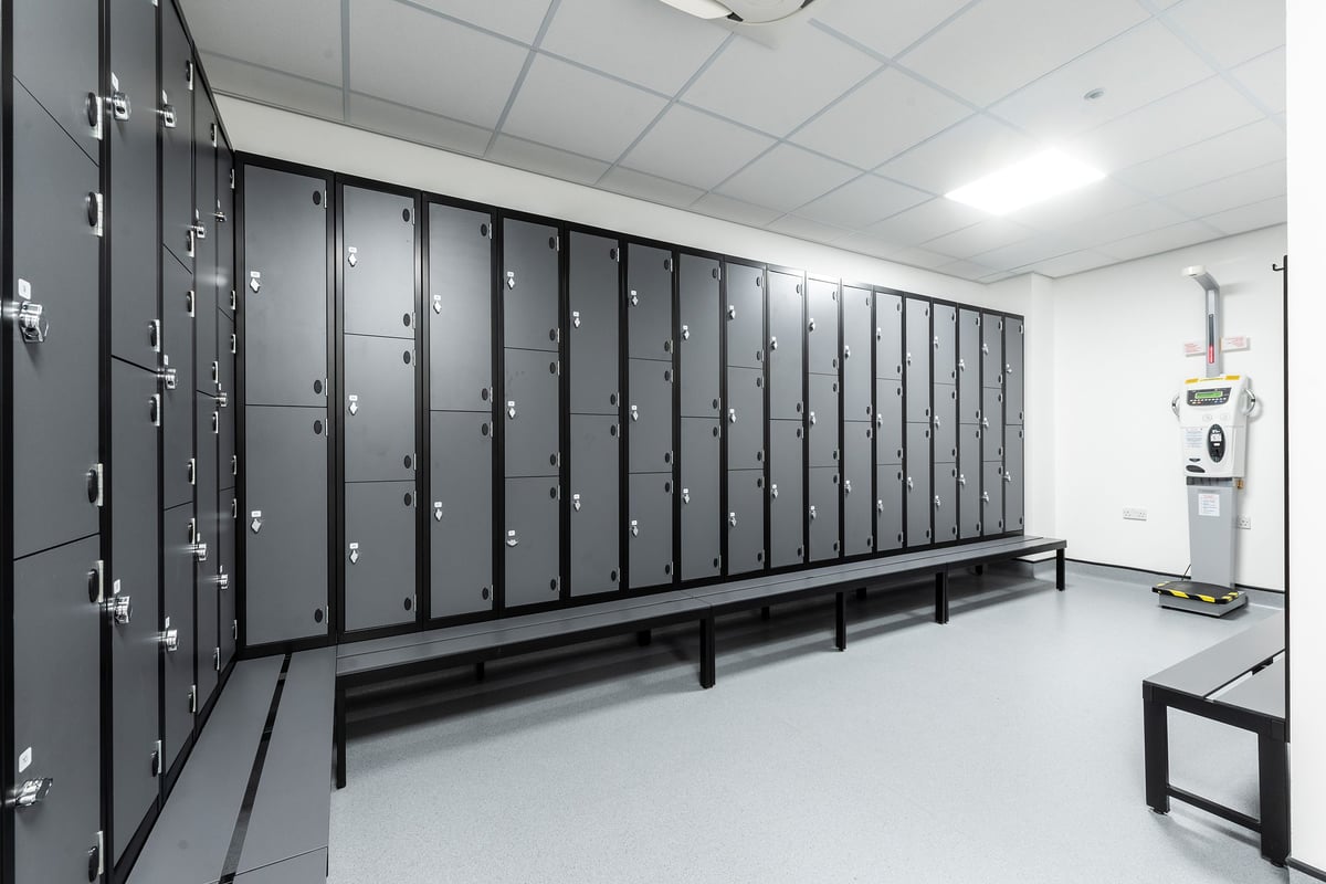 PureGym London Crouch End: Rows of lockers, benches, and an electronic kiosk in a bright, tiled room.