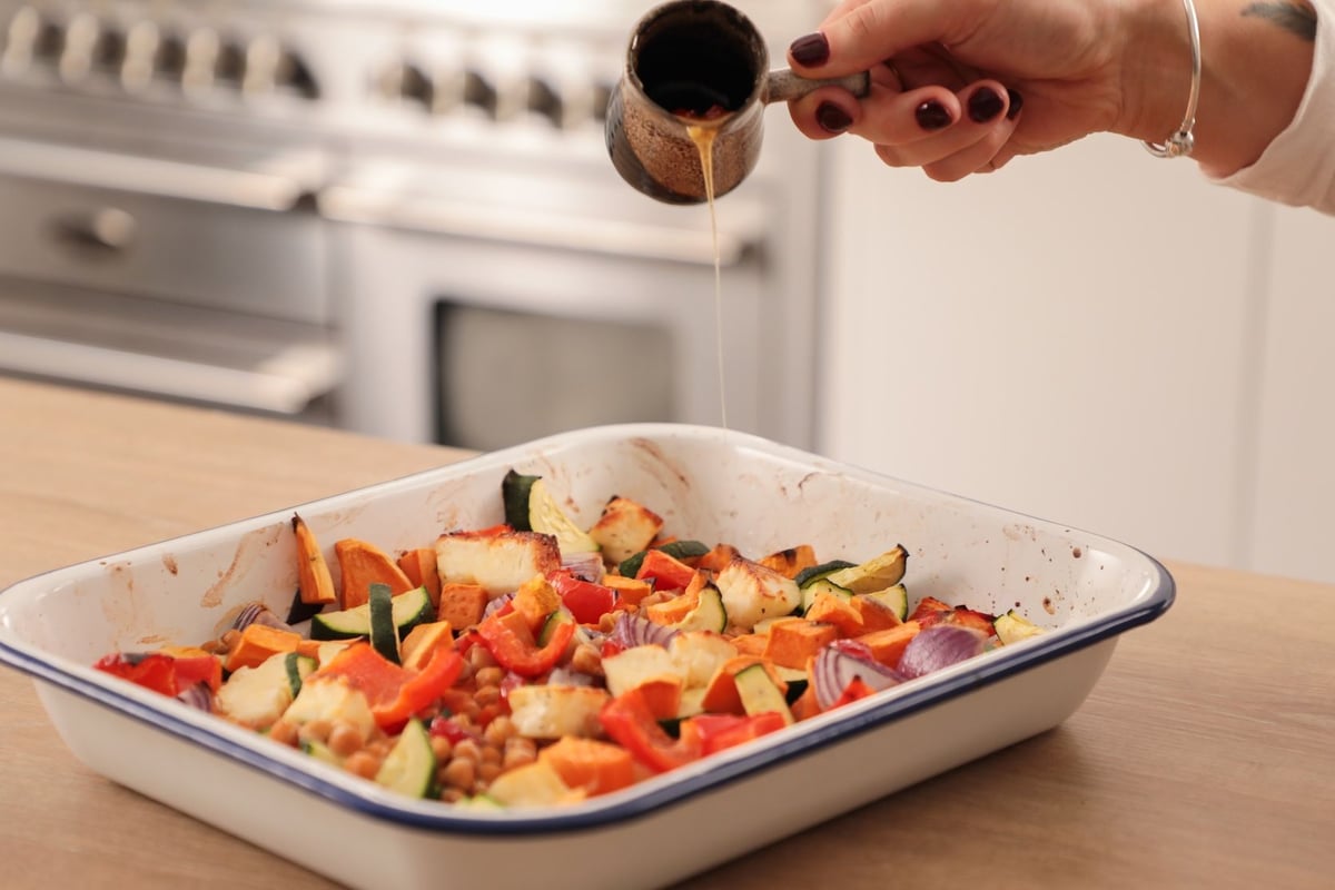 Hand drizzling sauce over roasted vegetables and halloumi in white enamel baking tray