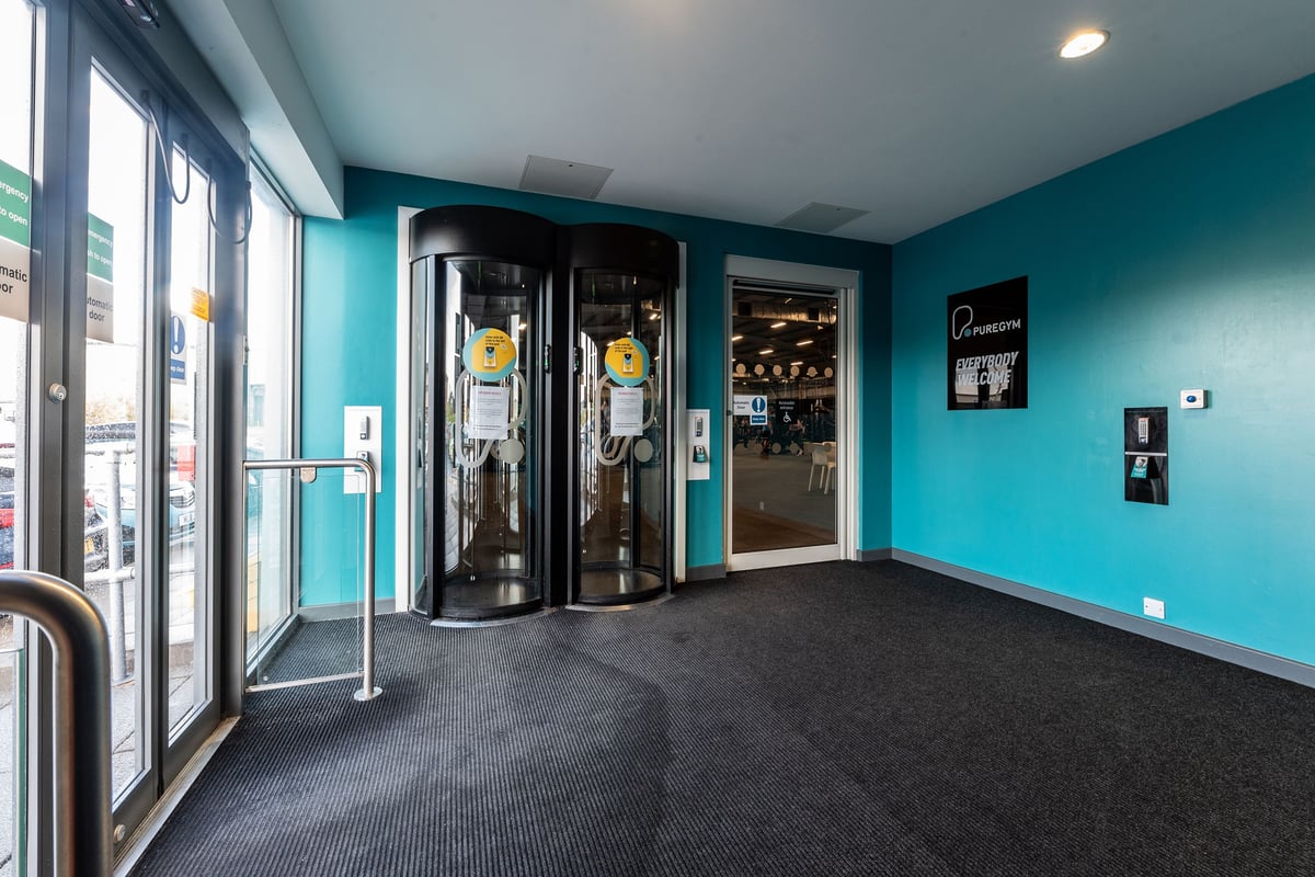 PureGym Widnes: Revolving door entry to fitness center, teal walls, dark carpet flooring.