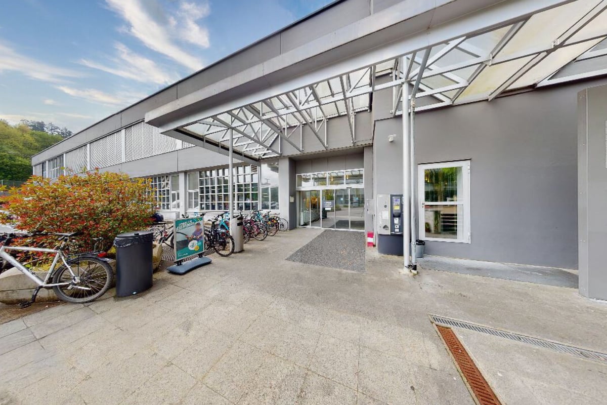 PureGym Bern Bümpliz: Extérieur du gymnase avec supports à vélos couverts, entrées et poubelles dans la zone pavée.