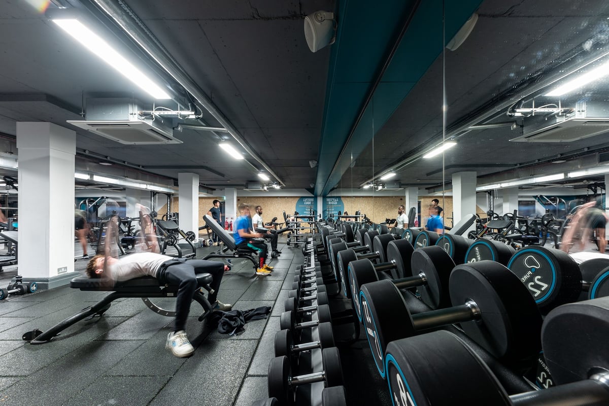 PureGym London Putney: Rows of stationary bikes, weight benches, dumbbells, and mirrors in a dimly lit gym interior.