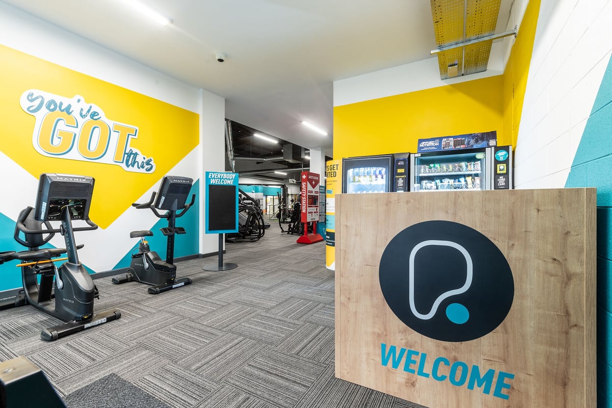 PureGym Dungannon: Exercise bikes, weights area, welcome sign, bright yellow and teal color scheme, open fitness space.