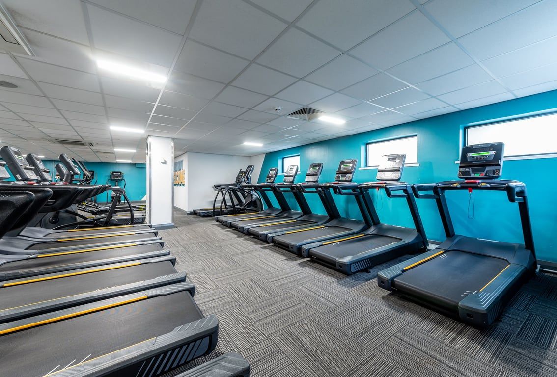 PureGym Northolt: Rows of treadmills in a teal and grey toned fitness centre with TVs mounted on walls.