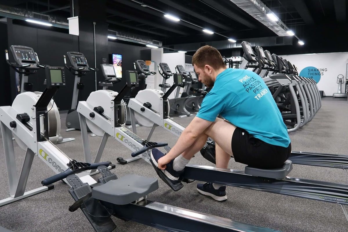 Man in blue shirt adjusting shoes on rowing machine in gym with cardio equipment
