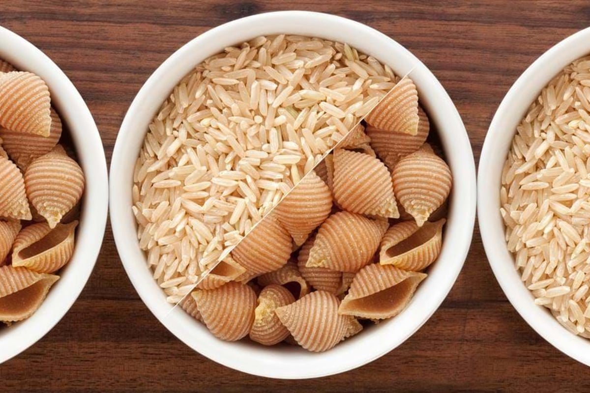 Three white bowls with brown rice and shell-shaped pasta on a wooden surface