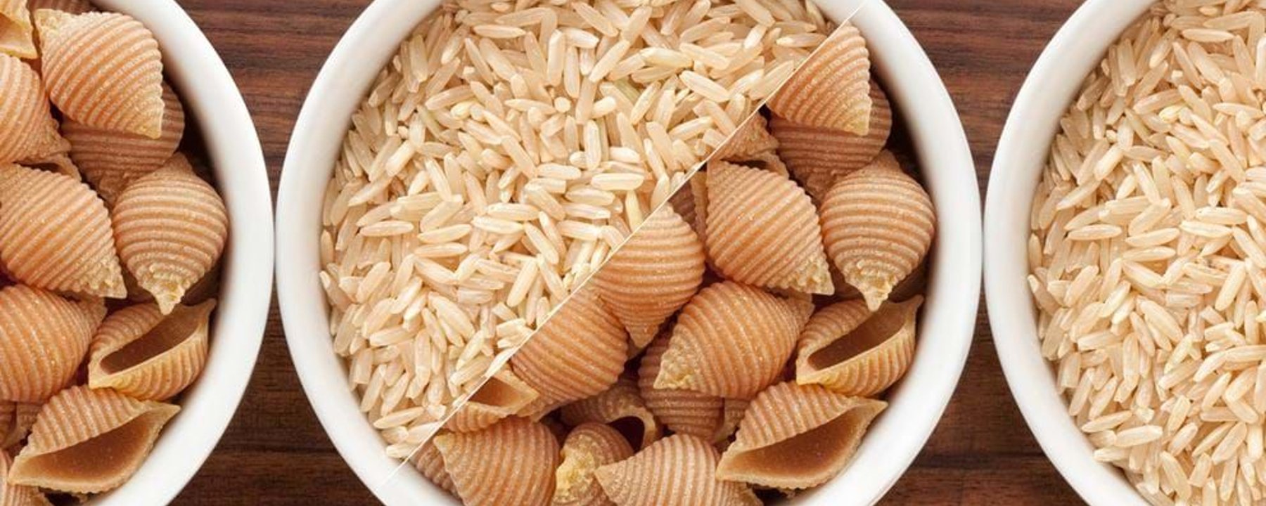 Three white bowls with brown rice and shell-shaped pasta on a wooden surface
