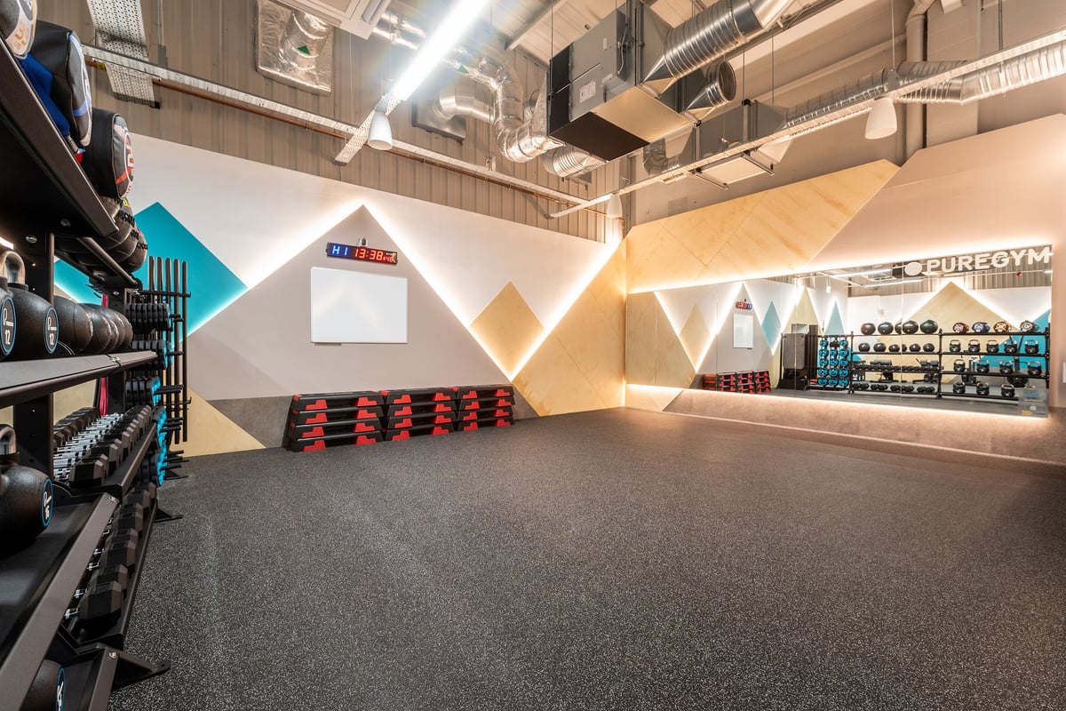 PureGym Alfreton: Weight racks, geometric wall designs, rubberised flooring, exposed ceiling ducts in a modern gym interior.