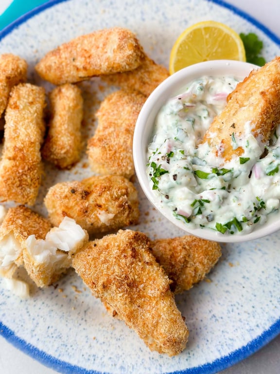 Breaded fish sticks with herb dip and lemon wedge on a speckled plate