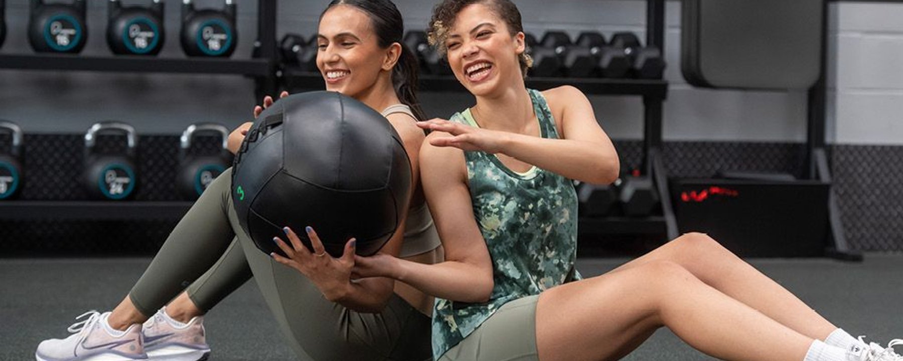 Two women sitting back to back passing a medicine ball in a gym.