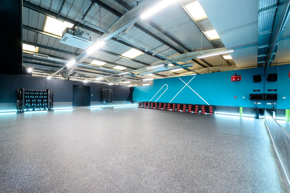 PureGym London Hayes: Gym studio with sled tracks, weight racks, and rubberized flooring under industrial ceiling.
