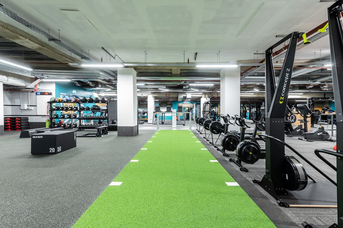 PureGym London Monument: Weightlifting area with dumbbells, resistance machines, green turf runway, and rubberized flooring.