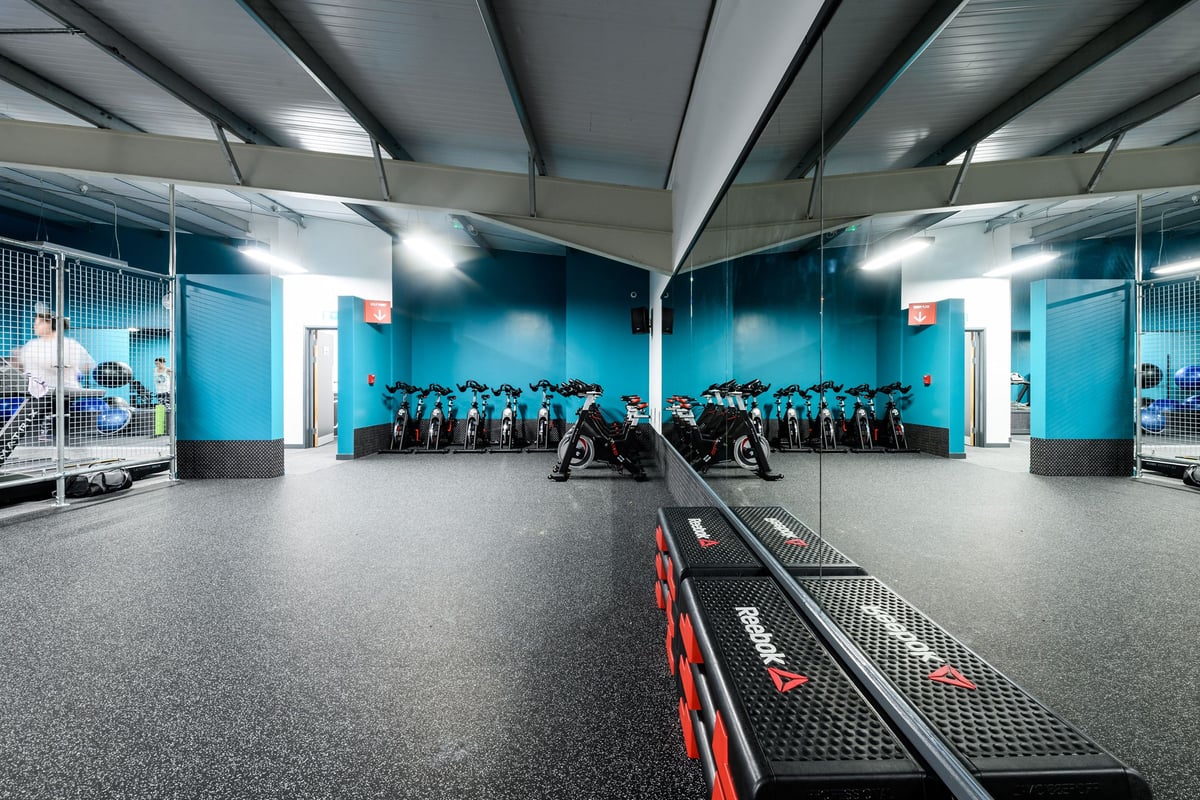 PureGym Swindon Mannington Retail Park: Rows of stationary bikes, weight racks, and floor mats in a teal-lit studio space.