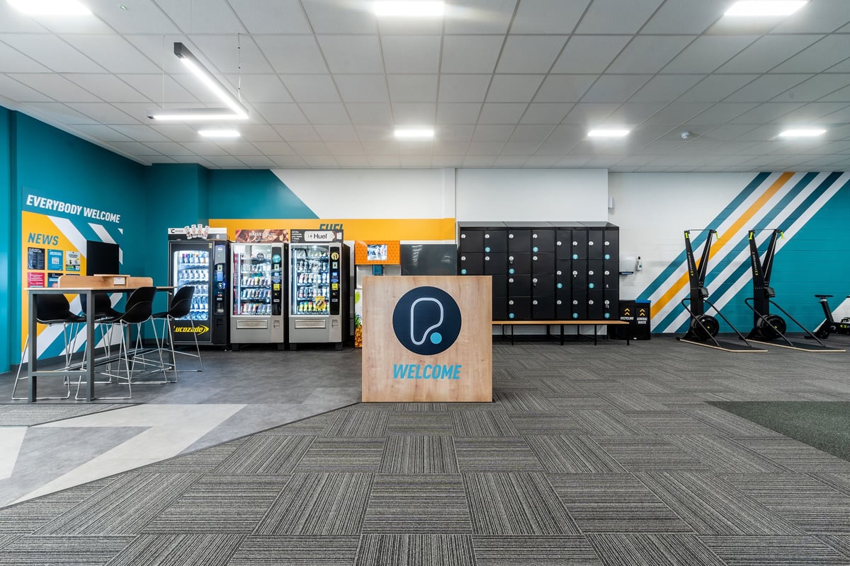 PureGym Coventry Warwickshire Shopping Park: Welcome area with vending machines, lockers, seating, and teal branding accents.