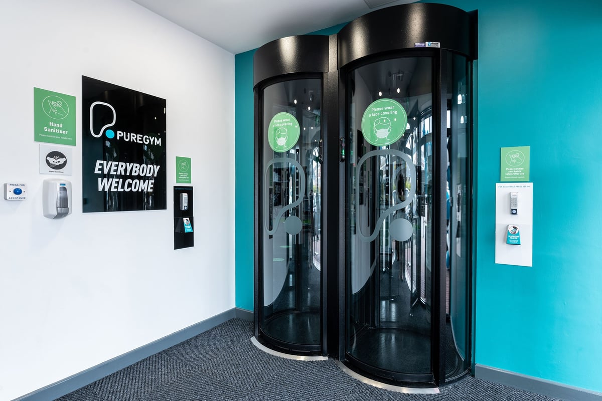 PureGym Warrington Central: Two glossy black cylinders for full-body sanitisation, "Everybody Welcome" signage on teal walls.