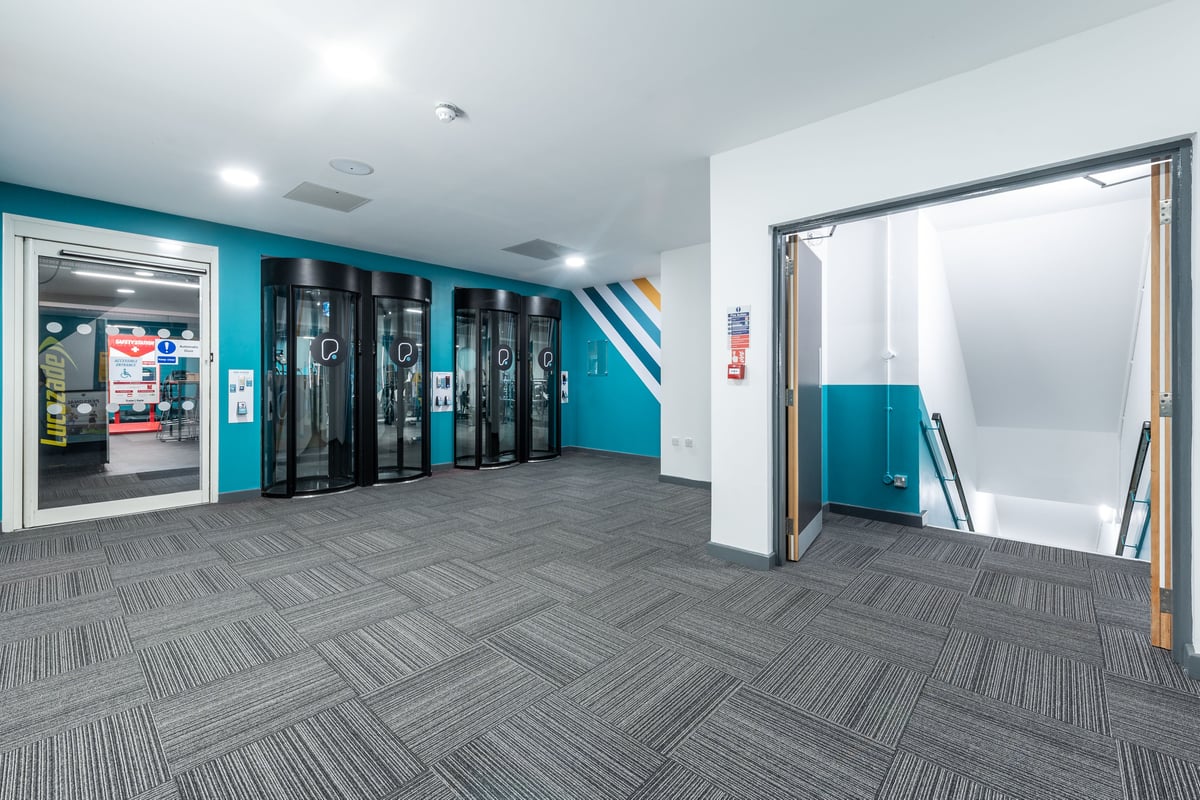 PureGym London Ilford: Entry area with turnstile gates, vending machines, teal accents, industrial carpet tiles.