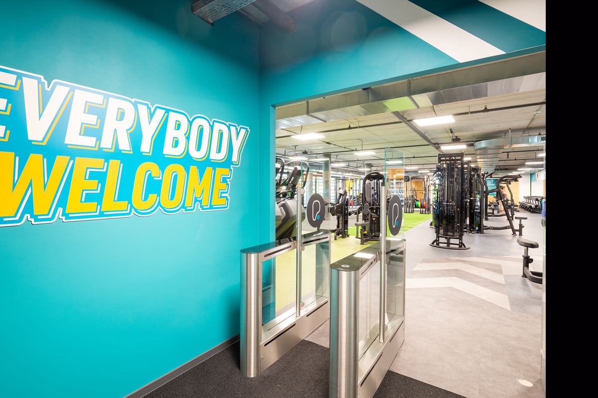 PureGym Genf Le Bouchet: Large gym area with weight machines, washing machines, and a bright "Everybody Welcome" sign.
