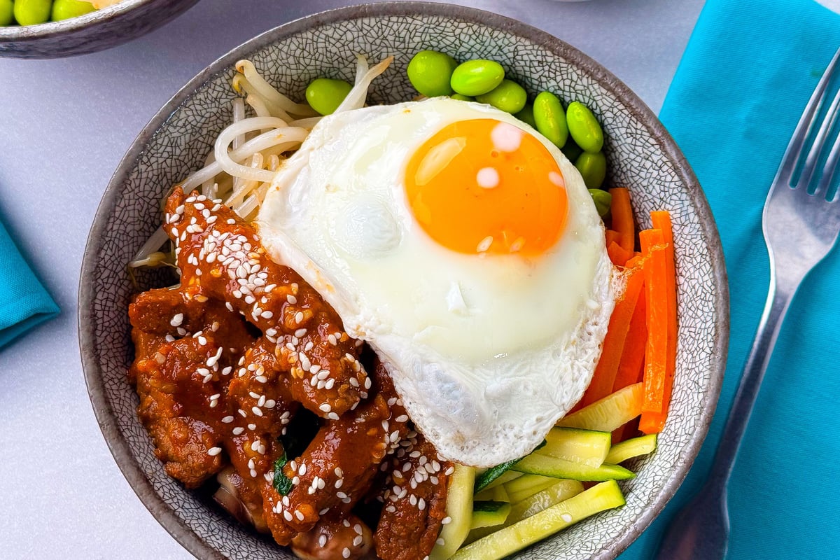 Bowl with fried egg, glazed meat with sesame seeds, edamame and sliced vegetables