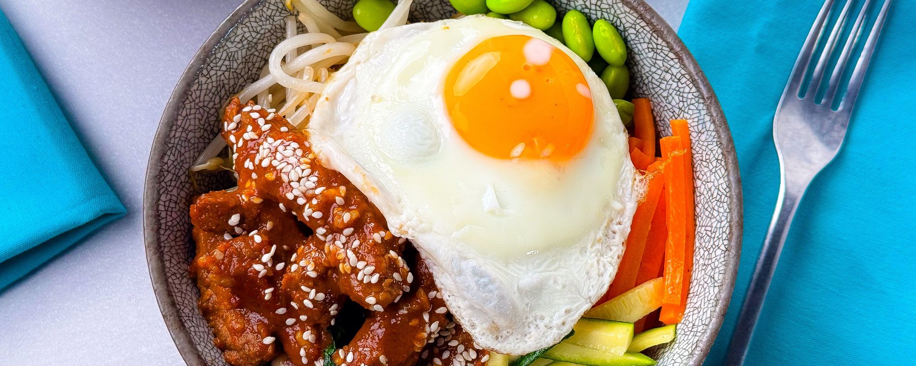 Bowl with fried egg, glazed meat with sesame seeds, edamame and sliced vegetables