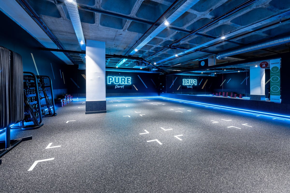 PureGym Maidstone The Mall: Rubberized flooring, weight stands, mirrors, and neon lighting in a dimly lit studio space.