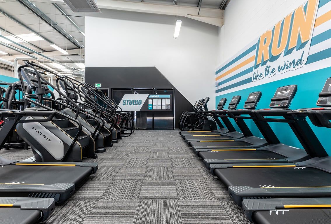 PureGym Dumfries: Rows of treadmills alongside graffiti wall with 'Run' logo, studio signage visible.