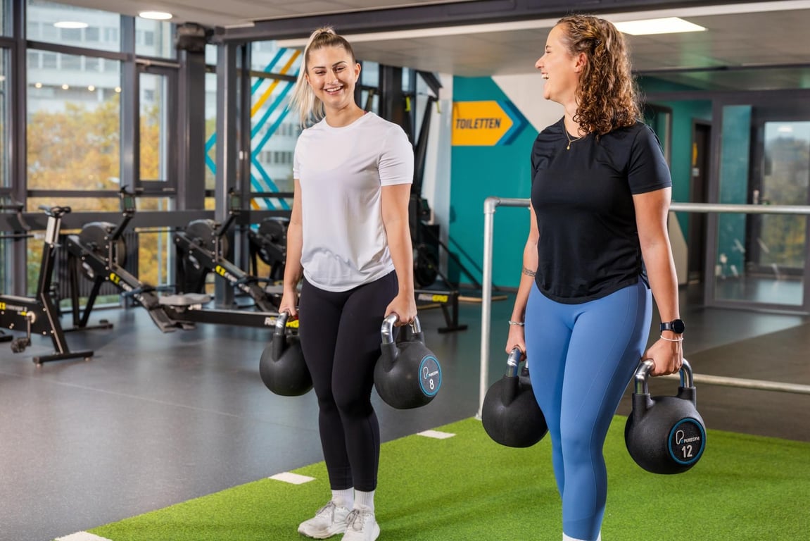 Zwei lächelnde Frauen tragen gemeinsam Kettlebells auf dem Trainingsboden im PureGym Zürich