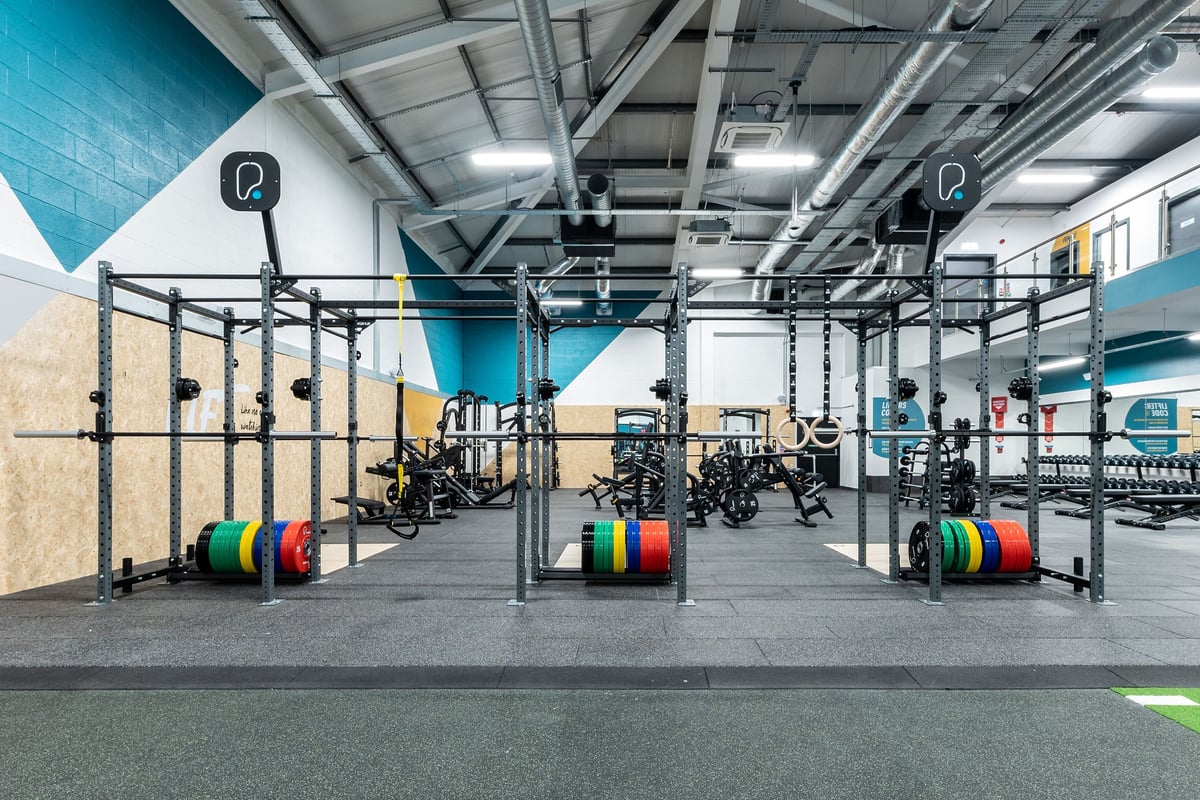 PureGym Ballymena: Squat racks, weight plates in multiple colours, rubberized flooring, and overhead rigging in a gym setting.