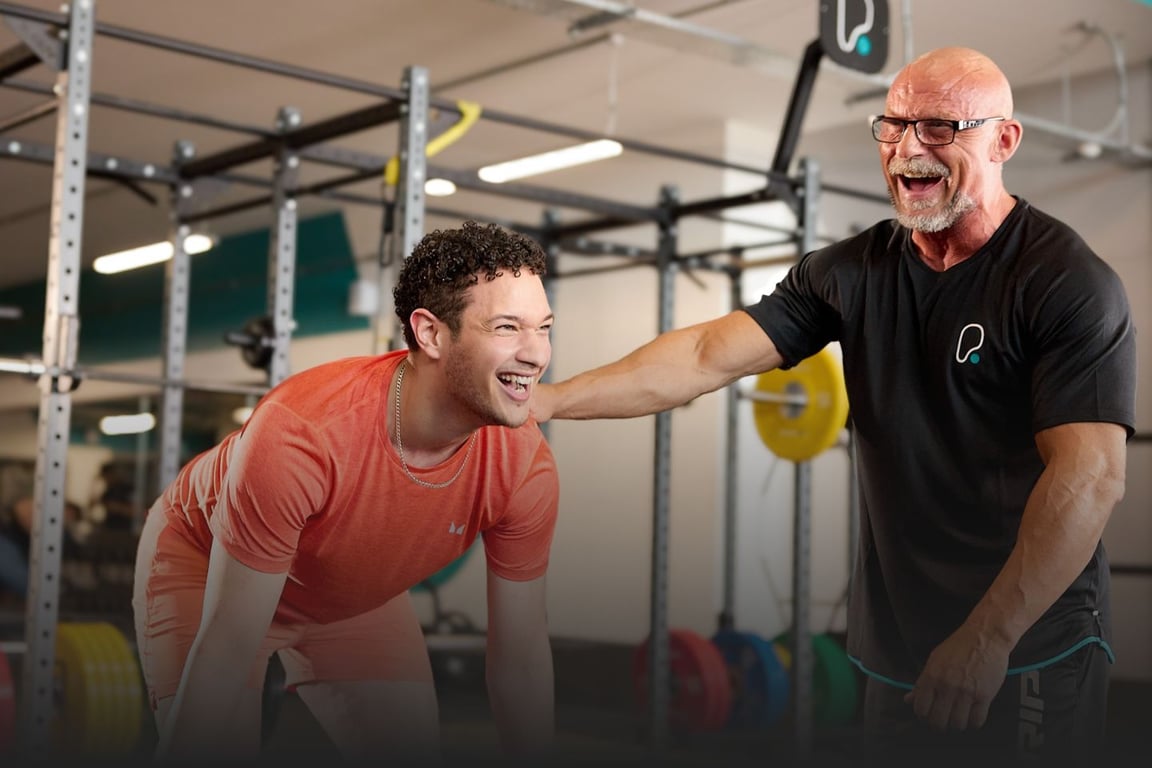A young, male PureGym member in orange sports gear performing a deadlift being encouraged by and older, male personal trainer wearing a PureGym-branded black top