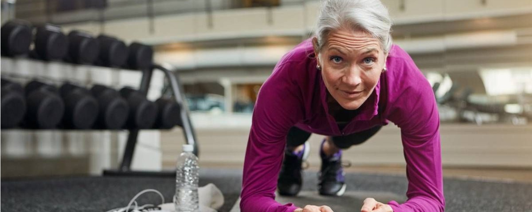Femme âgée en haut violet faisant la planche sur tapis dans une salle avec poids proches