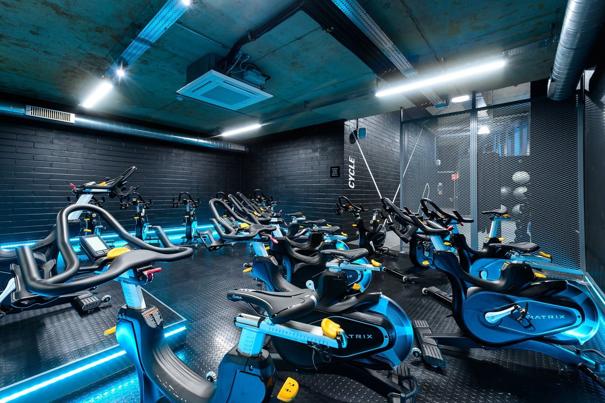 PureGym London Kidbrooke Village: Studio with rows of stationary bikes, neon lighting, and exposed ductwork on ceiling.
