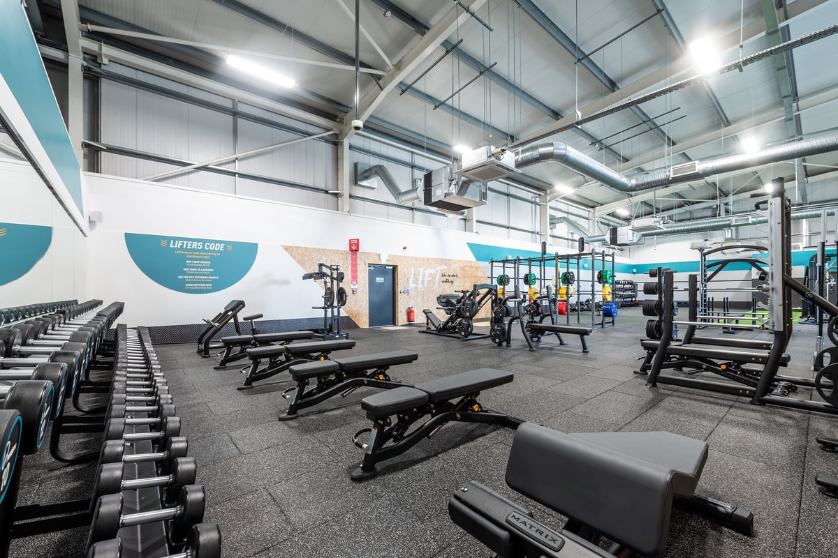 PureGym Stamford: Weightlifting area with benches, dumbbells, racks, rubberized floors, and exposed ceiling beams.